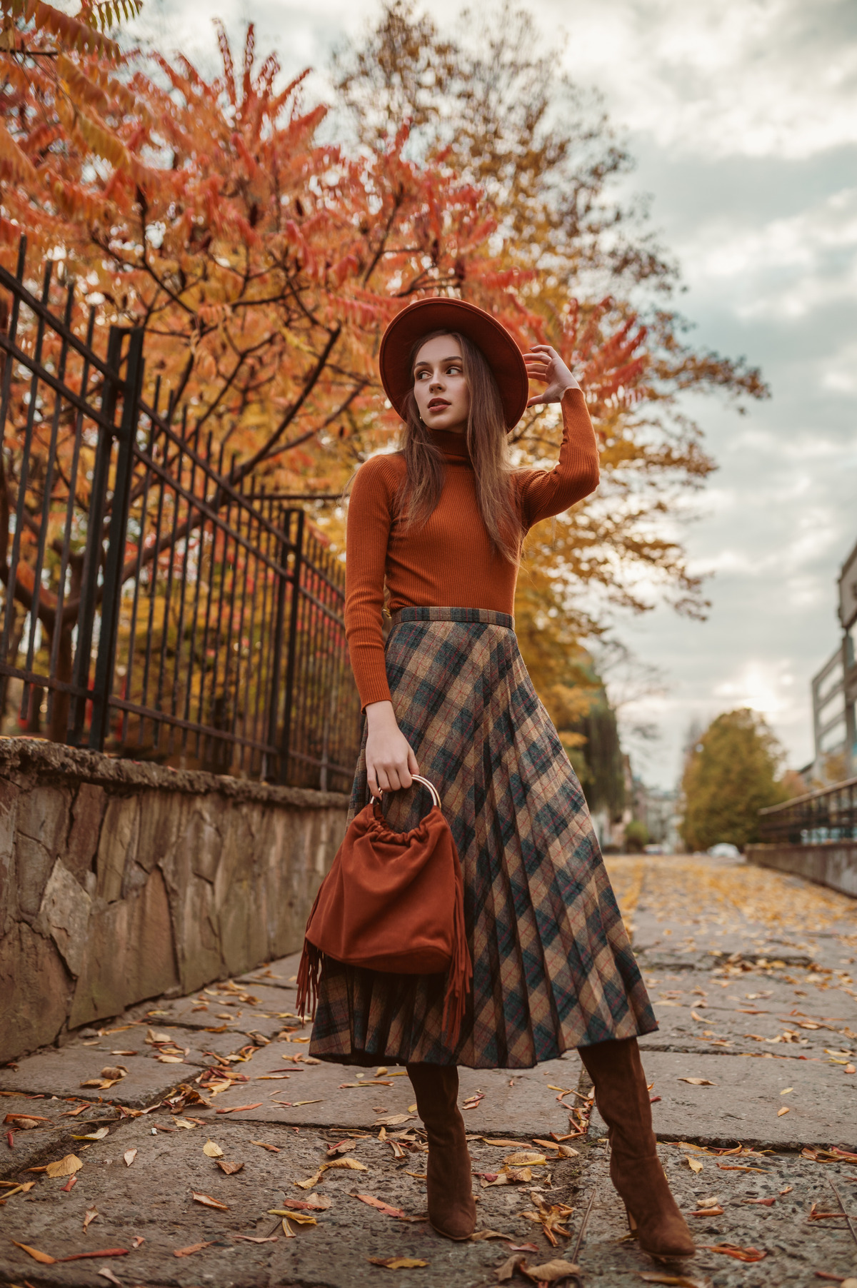 elegant woman wearing stylish orange hat, turtleneck, checkered midi skirt, high boots holding suede bag with fringe, walking in street of city